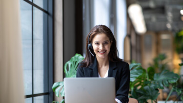 Mitarbeiterin mit Headset sitzt lächelnd neben einem großen Fenster im Büro an einem digitalen Arbeitsplatz.