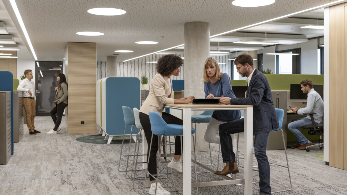 In einem modernen Büroraum arbeiten drei Personen zusammen an einem Stehtisch, zwei weitere Personen unterhalten sich im Hintergrund.