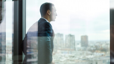 Mann im Anzug schaut aus einem Bürofenster auf die Stadt. Im Vordergrund eine leicht reflektierende Fensterscheibe.